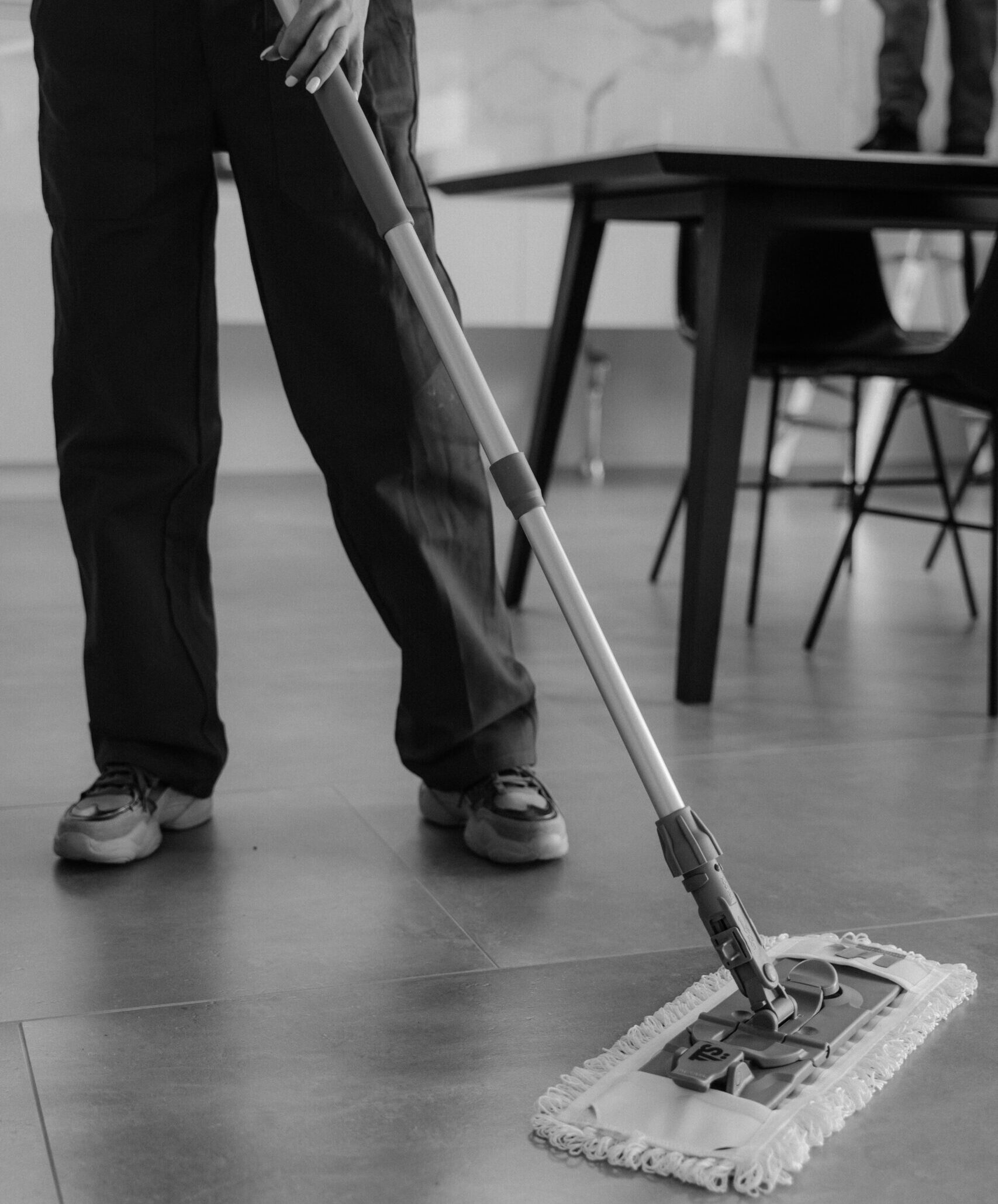 Monochrome image of a person mopping a tiled floor in an indoor setting.